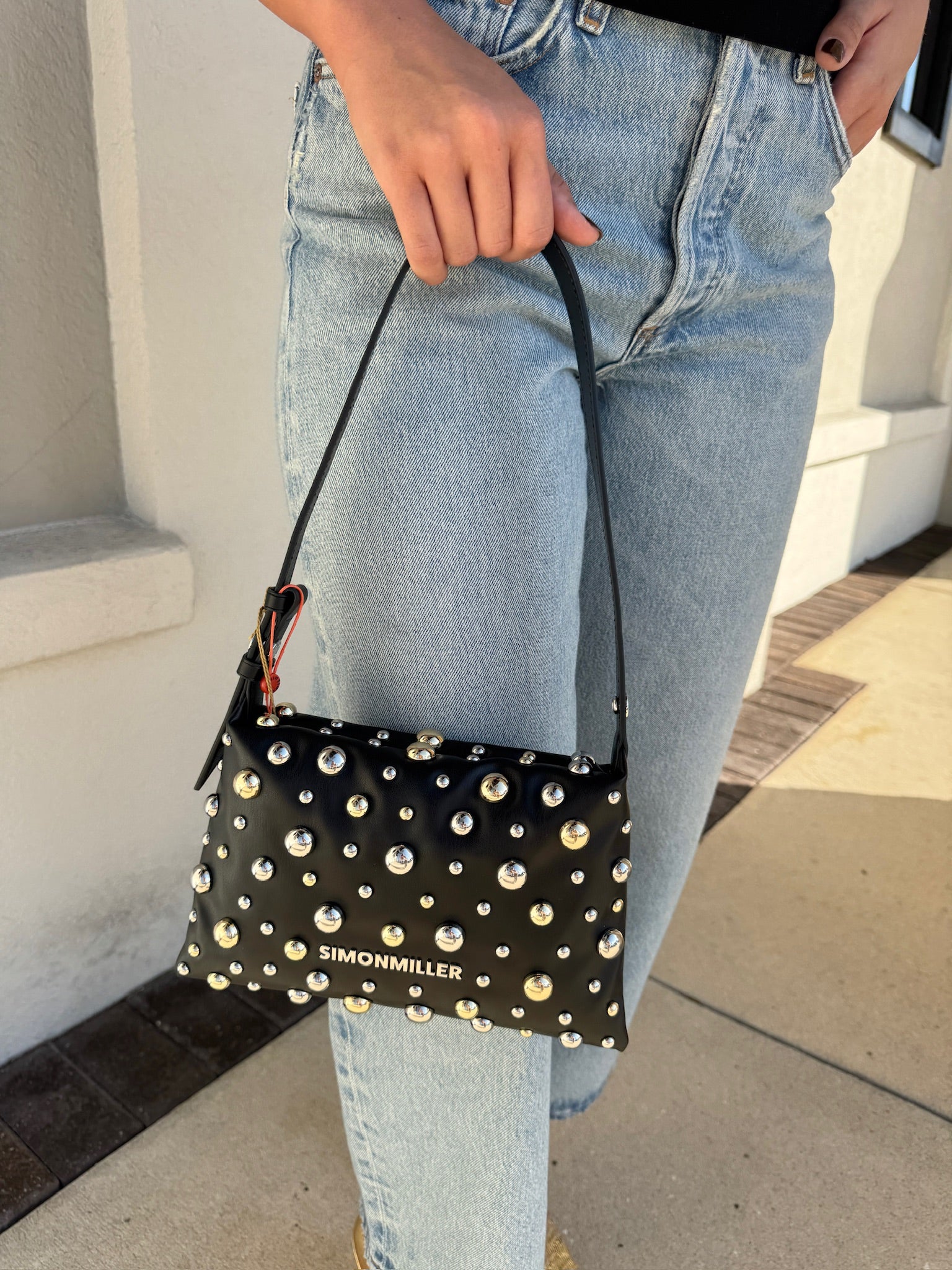Person holding a black clutch with gold studs and 'Simon Miller' branding.