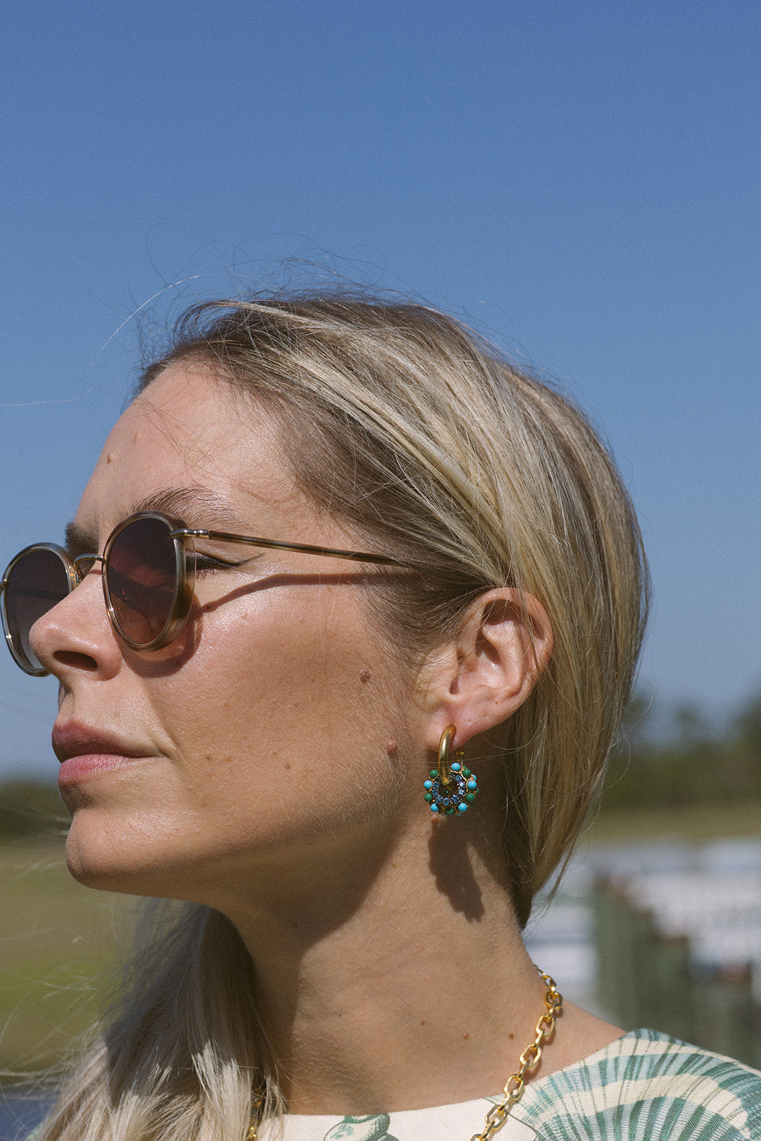 Woman wearing sunglasses and a gold necklace with a blue earring against a clear blue sky.