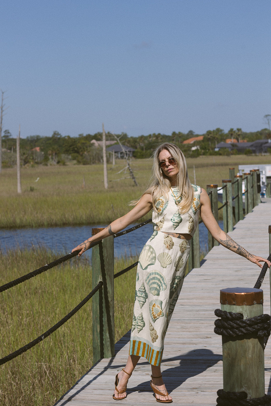 Woman in a patterned set standing on a wooden dock by a body of water with grass and trees in the background.