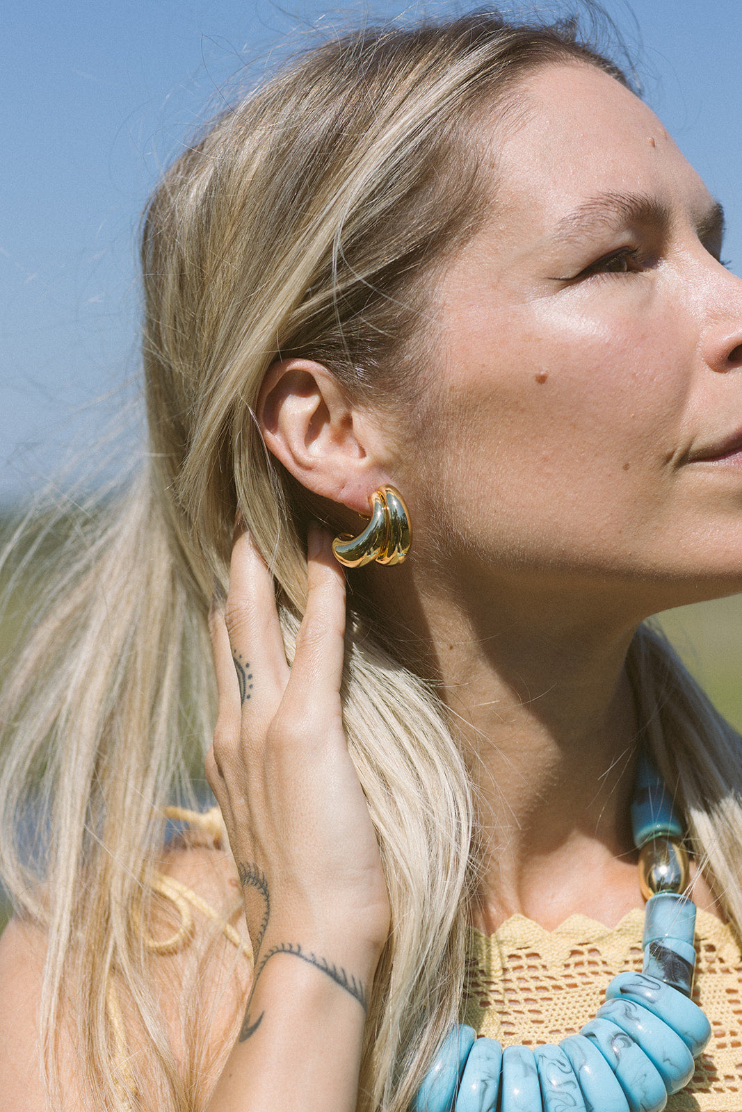 Woman wearing gold hoop earrings with a blue necklace against a clear sky.