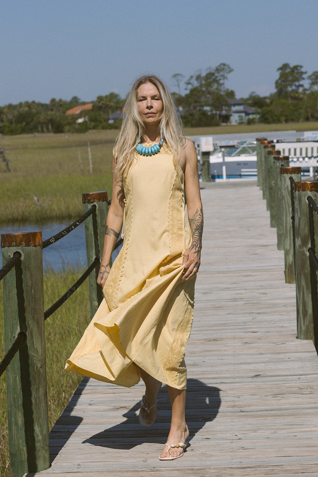 Woman in a yellow dress and Cely Sandals standing on a wooden boardwalk with a scenic background