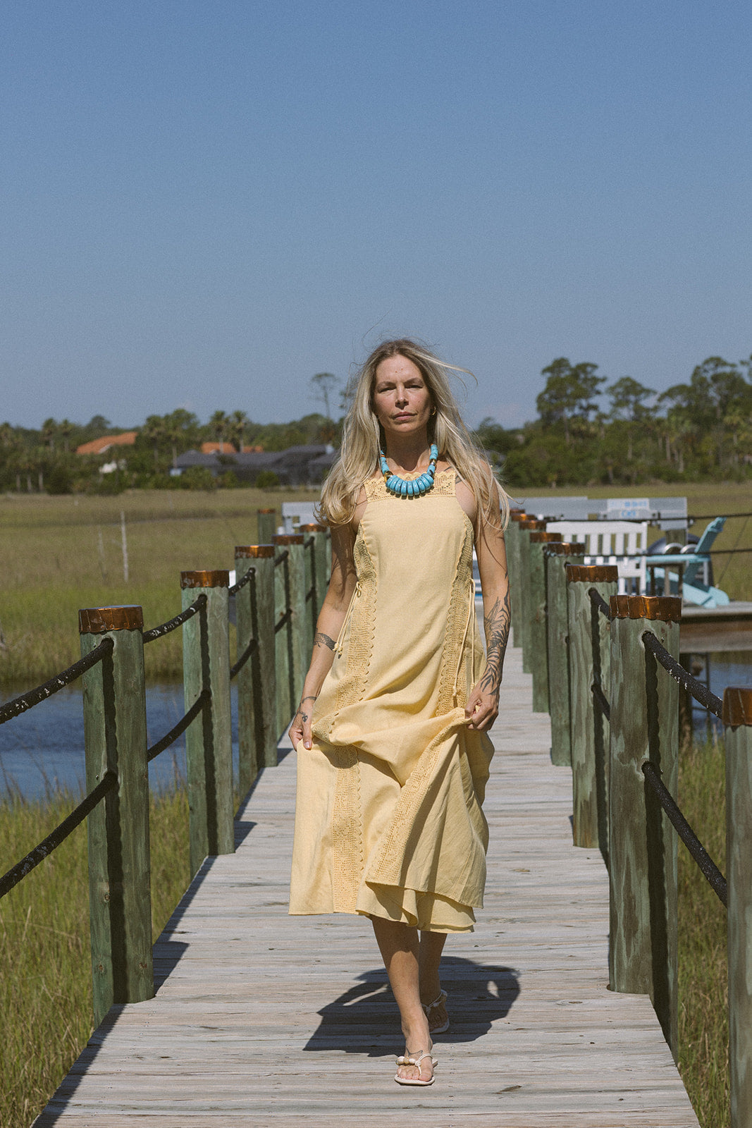 Woman in a yellow dress standing on a wooden boardwalk by a body of water with greenery in the background.