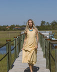 Woman in a yellow dress standing on a wooden boardwalk by a body of water with greenery in the background.