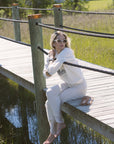 Woman sitting on a wooden dock by a body of water, wearing sunglasses and a white outfit.