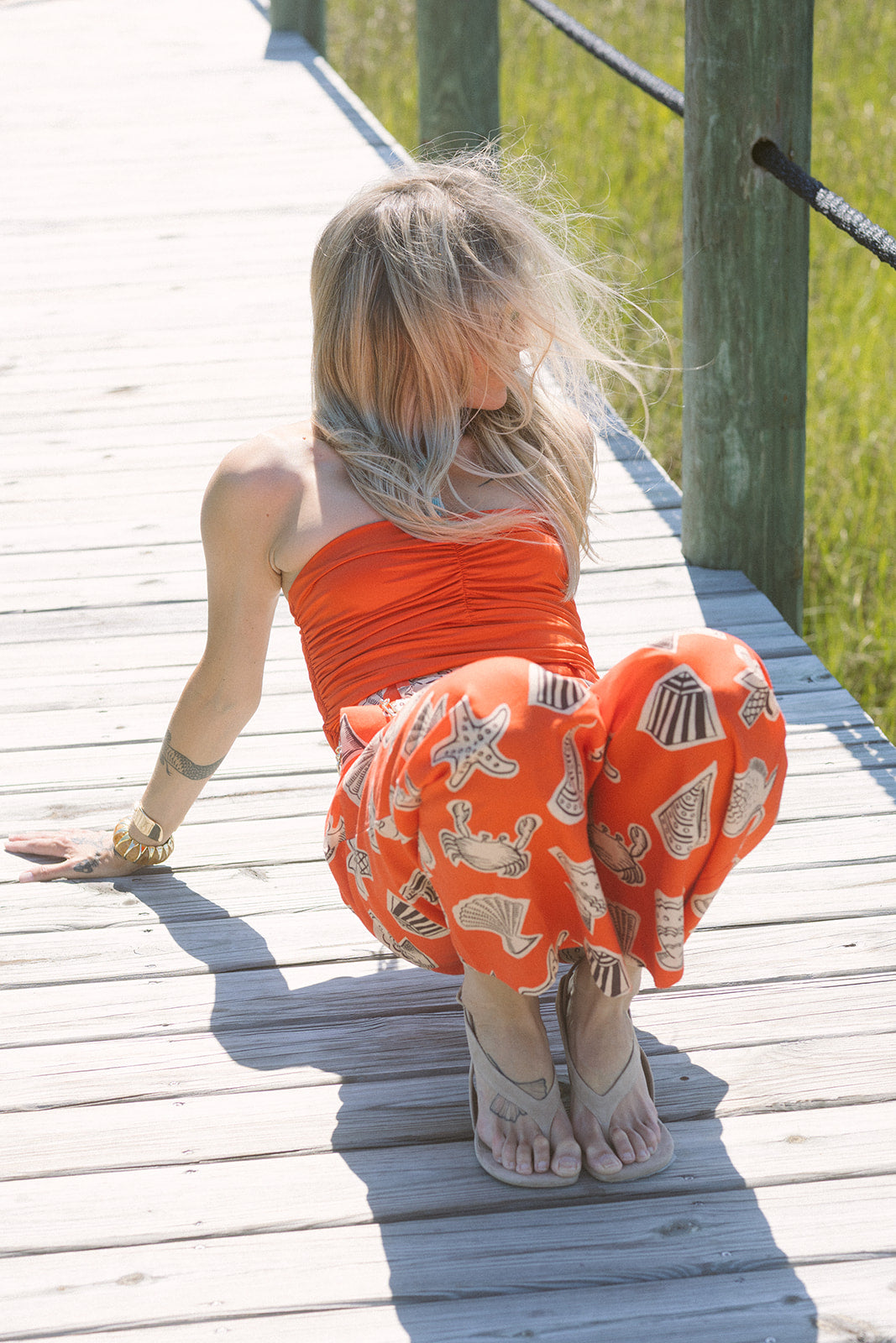 Woman sitting on a wooden dock wearing an orange outfit with Darya Suede Wedge Thong Sandal