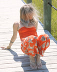 Woman sitting on a wooden dock wearing an orange outfit with Darya Suede Wedge Thong Sandal