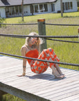 Woman in an orange outfit sitting on a wooden dock with a house and grass in the background