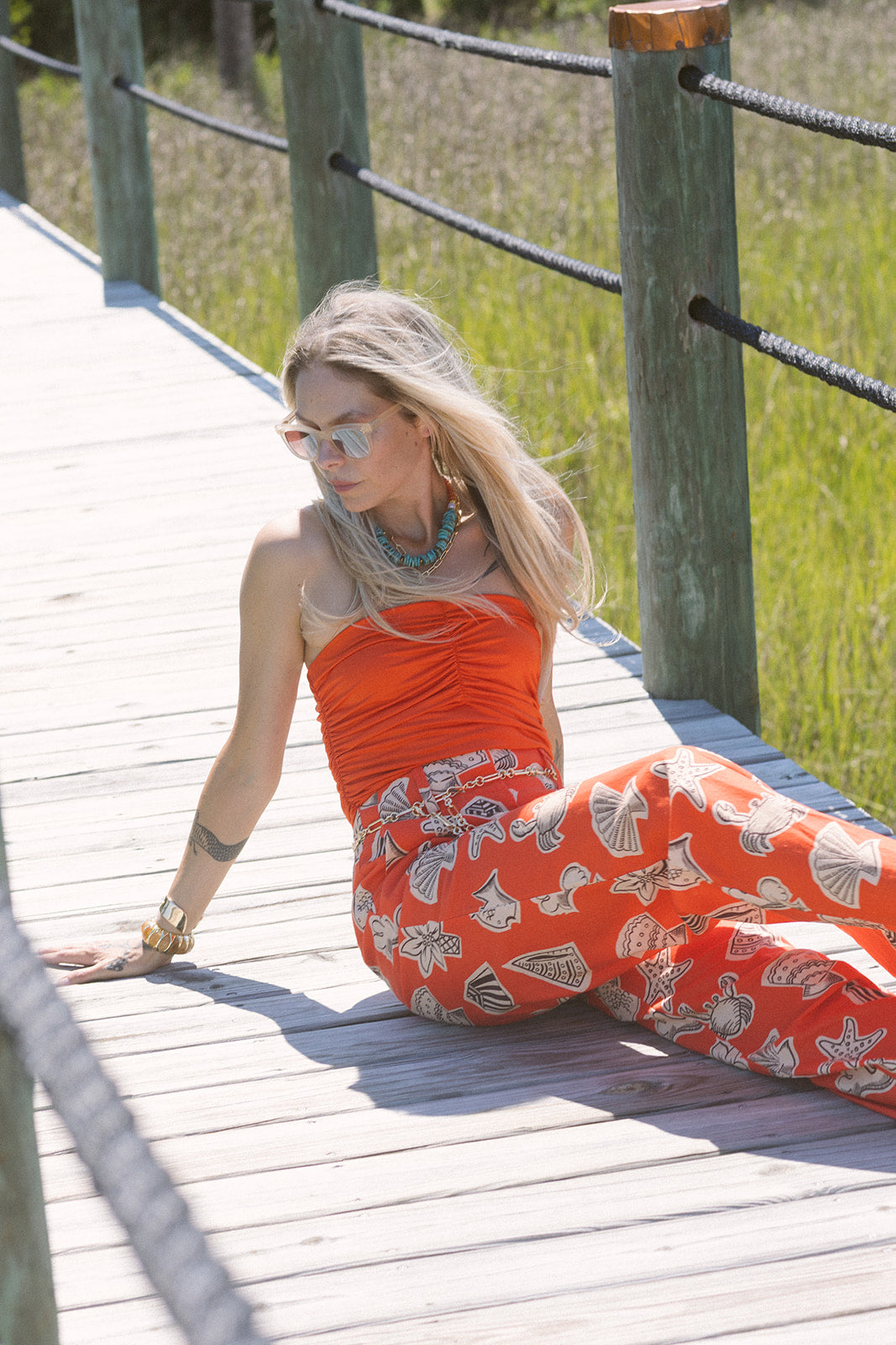 Woman in Tracey Necklace and an orange top and pants with a pattern sitting on a wooden bridge.