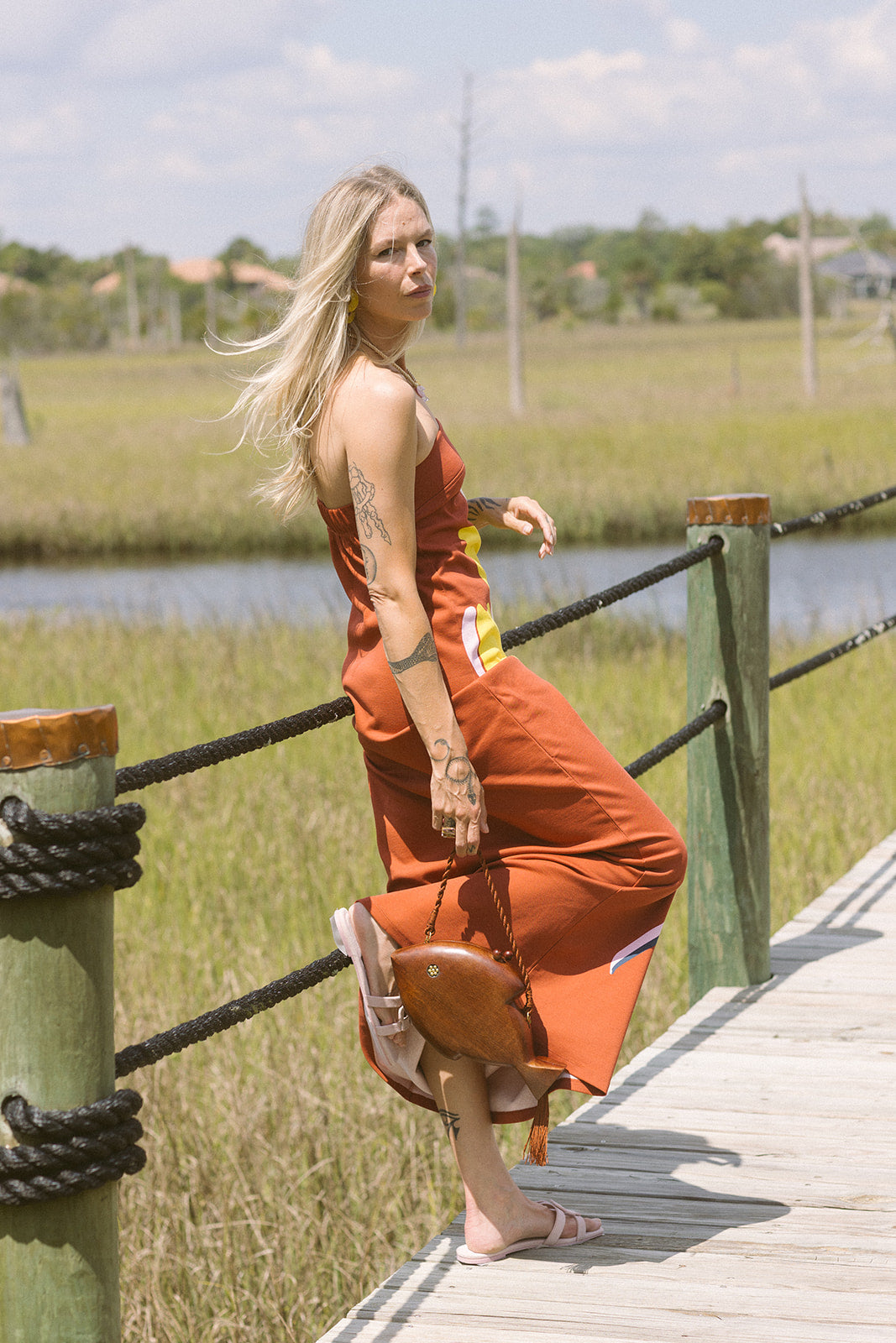 Woman in an orange dress and Monogram Suede Flat Slide standing on a wooden dock with grassy fields and water in the background.