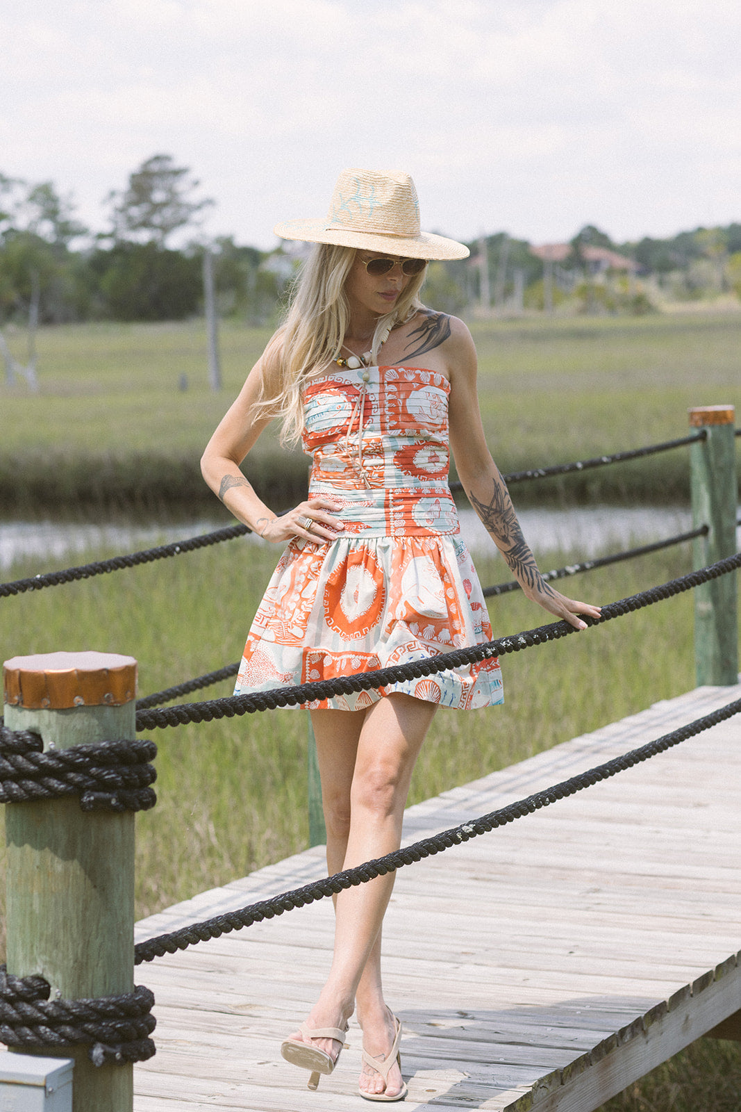 Woman in a patterned dress and hat standing on a wooden dock by a body of water. full body view
