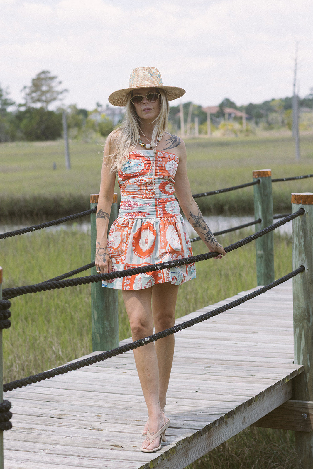 Woman in a patterned dress and hat standing on a wooden bridge over a waterway.