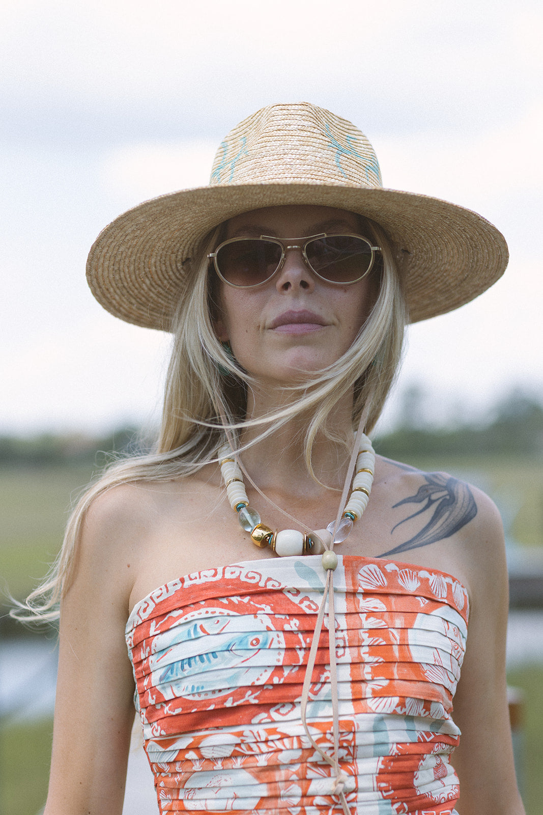 Woman wearing a straw hat and sunglasses with Coquina Necklace and a patterned dress outdoors.