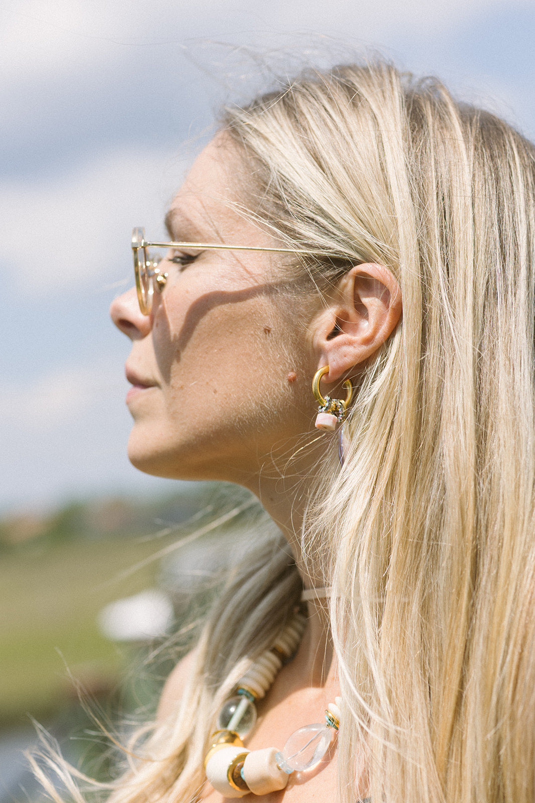 Close-up of a woman with blonde hair wearing gold earrings and sunglasses, outdoors.