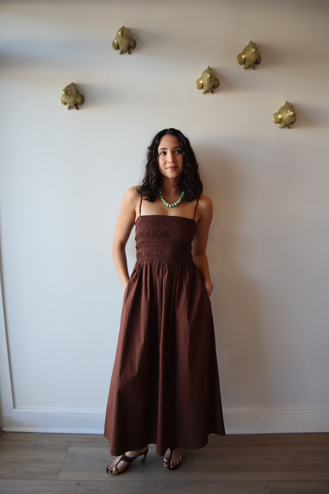 Woman in a brown dress standing against a white wall with gold decorative elements.