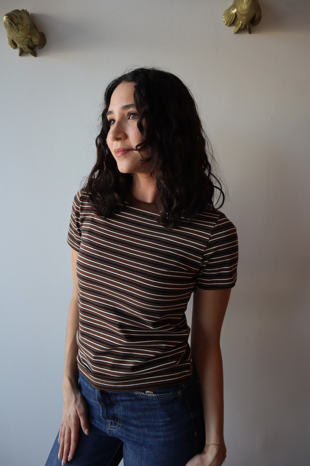 Woman in a striped shirt and jeans standing against a plain wall.