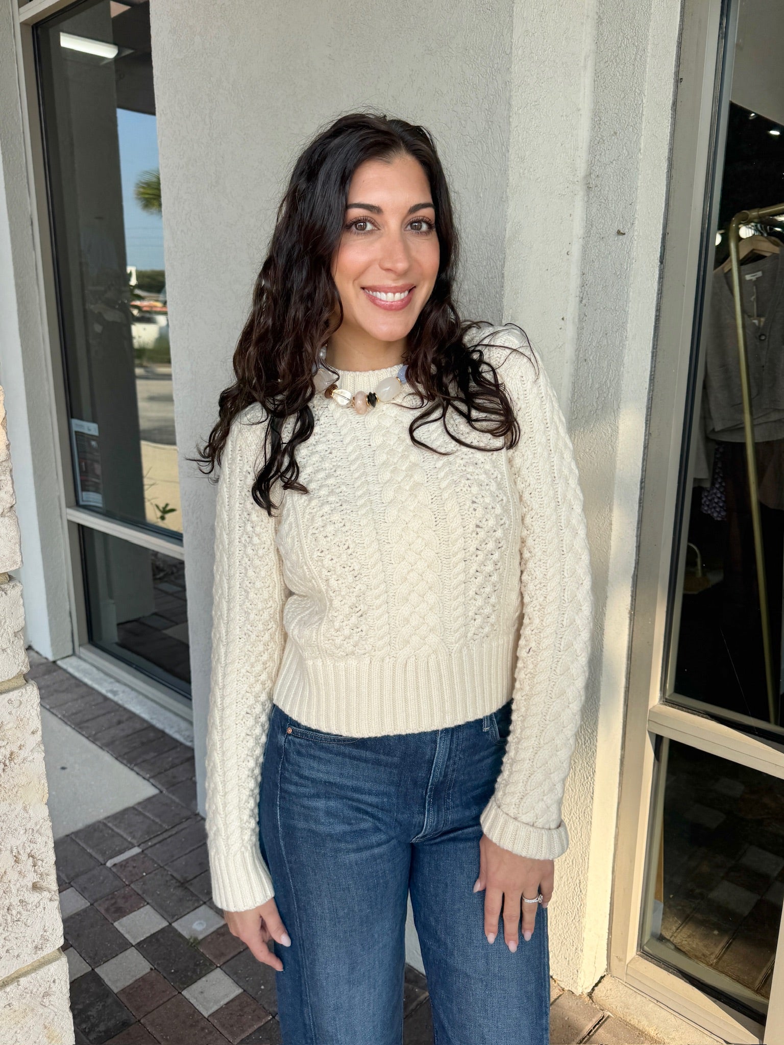 Woman wearing a cream sweater and blue jeans standing outside a building.