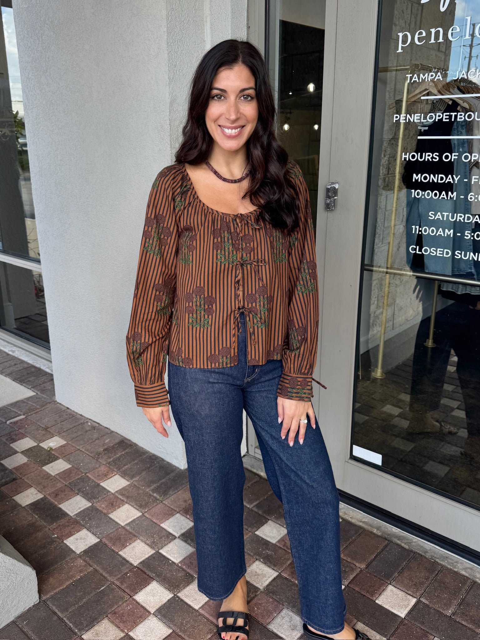Woman wearing a patterned blouse and jeans standing outside a store.