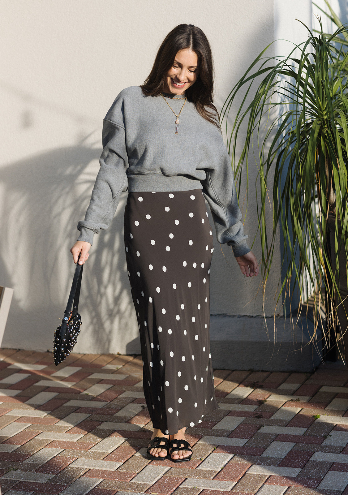 Woman in gray sweater and polka dot skirt standing outdoors with a plant in the background