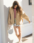 Woman in beige jacket and striped shirt walking outdoors on a sunny day.