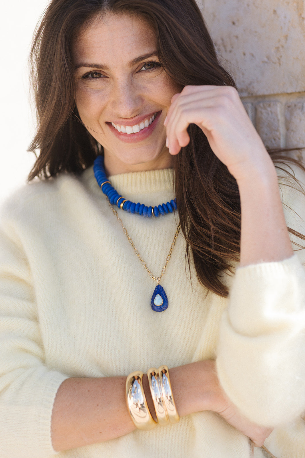 Woman wearing a white sweater, blue beaded necklace, and gold bracelets with a neutral background