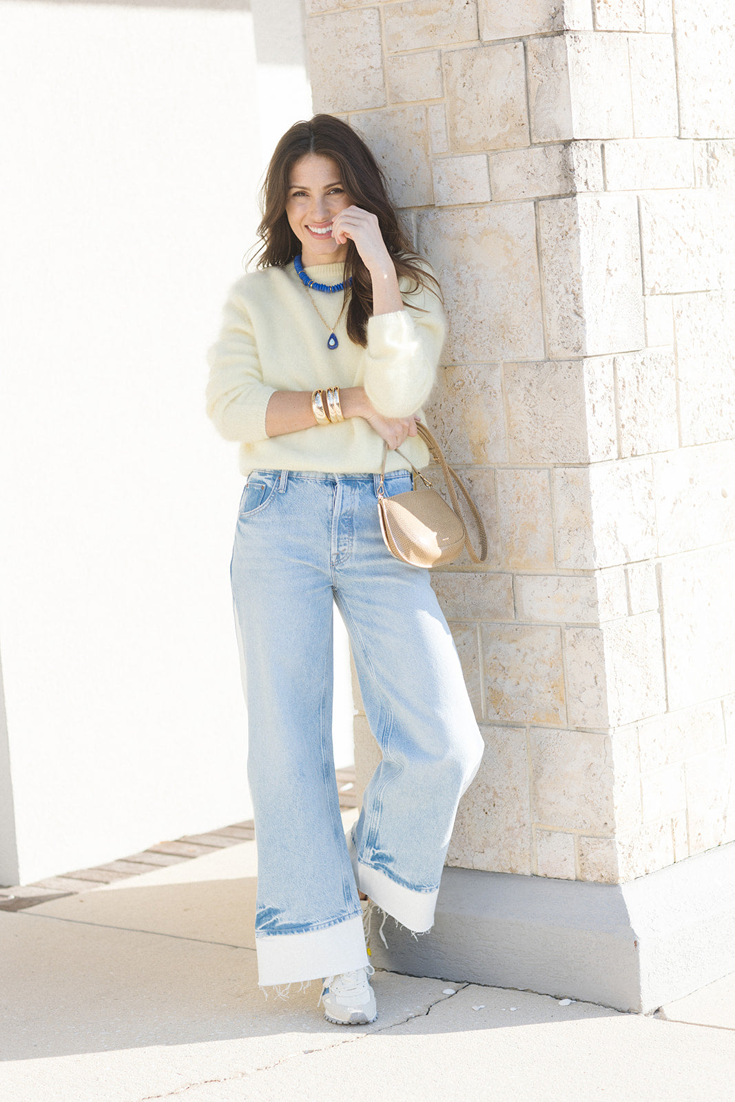 Woman in light sweater and jeans standing against a stone wall.