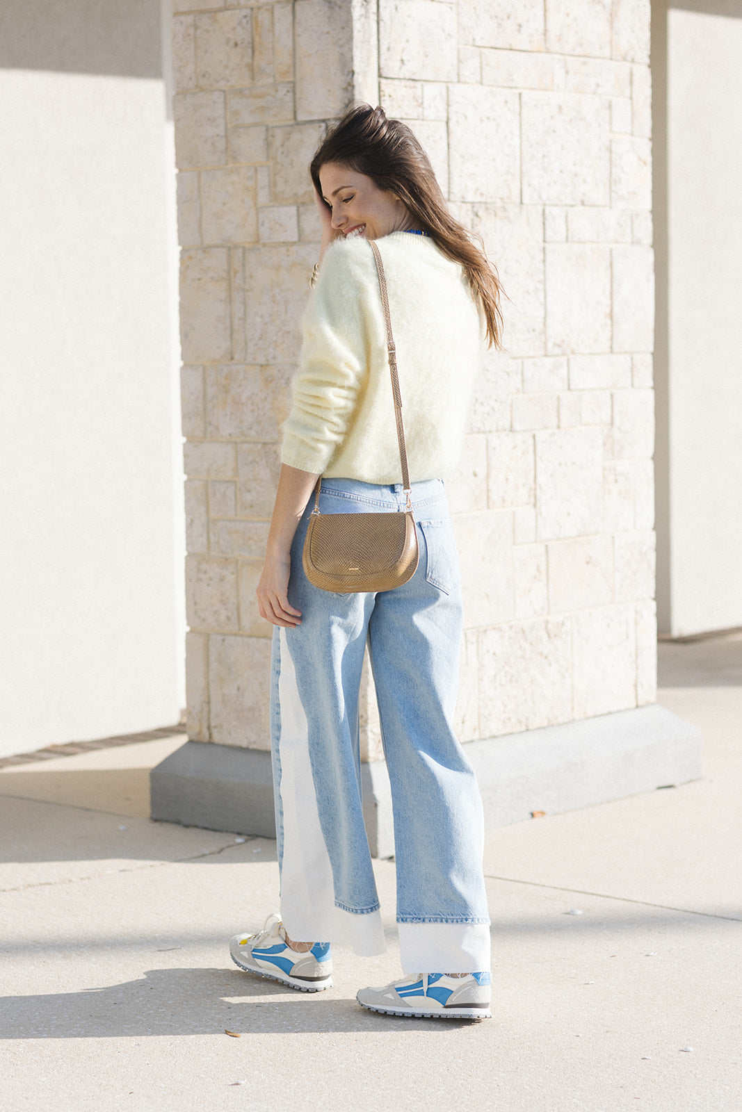 Woman walking outdoors wearing a light-colored sweater, blue jeans, and sneakers with a beige handbag.