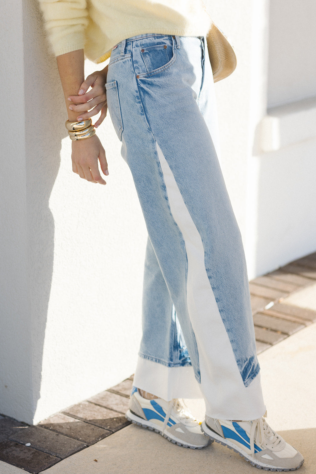 Person wearing light blue jeans with a white stripe, beige sweater, and sneakers on a wooden deck.