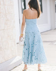 A woman viewed from behind wearing a strapless light blue midi dress with a scalloped lace hem, holding a white textured clutch on a paved walkway next to a light stone wall.