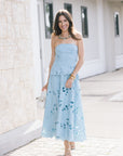 A woman wearing a light blue strapless lace dress with a peplum waist and flared skirt, standing outdoors on a paved walkway next to a light-colored building.