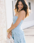 A woman wearing a light blue strapless dress with a textured, layered ruffle pattern, holding a white textured clutch bag. She is standing outdoors next to a light-colored stone wall.