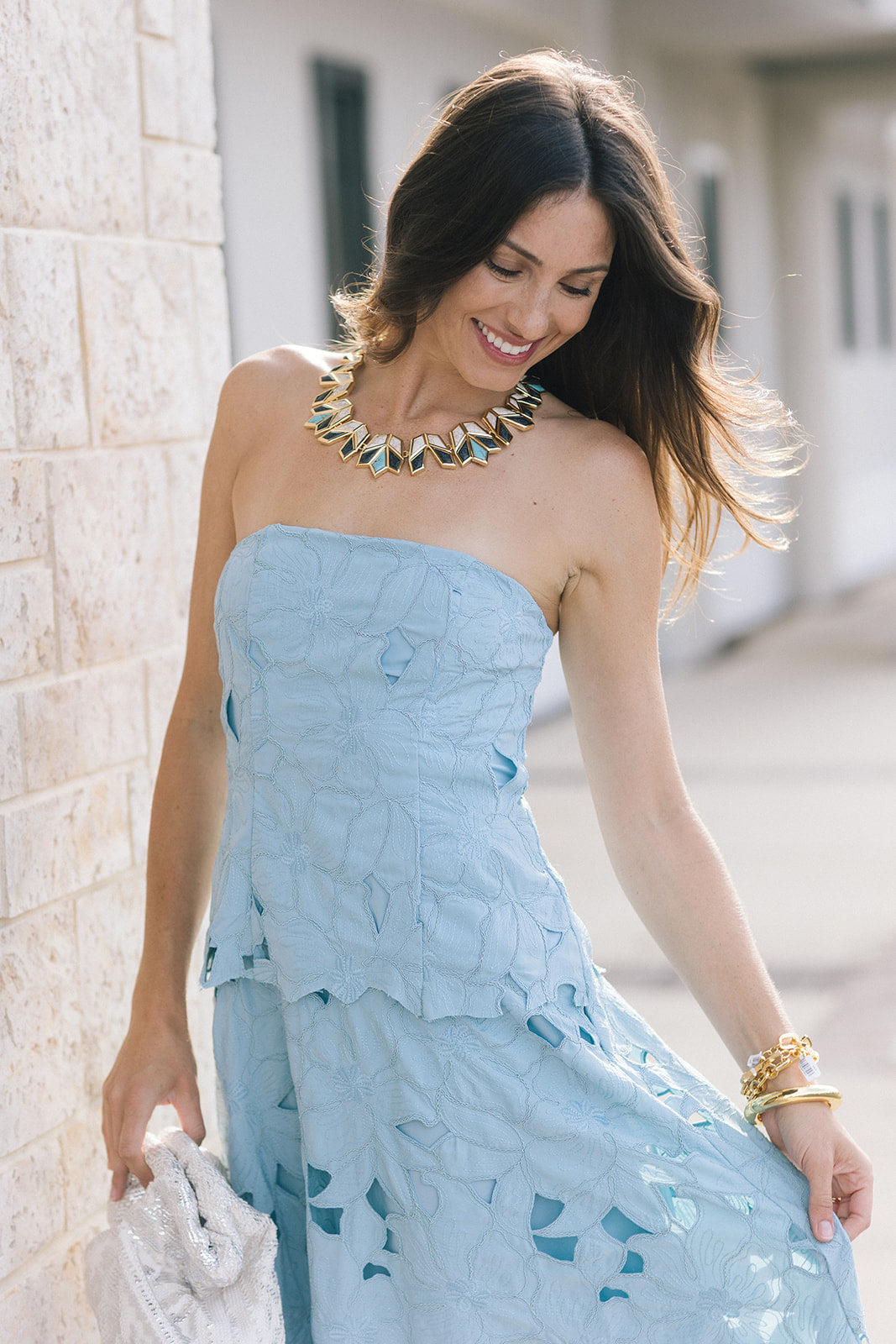 A woman wearing a light blue strapless dress with a textured, leaf-like pattern and cutout details, accessorized with a statement necklace and gold bracelets, standing outdoors next to a light stone wall.