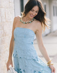 A woman wearing a light blue strapless dress with a textured, leaf-like pattern and cutout details, accessorized with a statement necklace and gold bracelets, standing outdoors next to a light stone wall.
