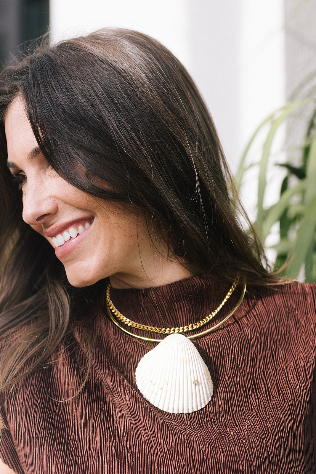 A woman with long dark brown hair smiling, wearinga large white seashell pendant necklace and a smaller gold chain.