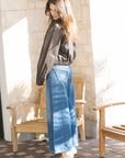 Woman wearing a gray blouse and blue jeans standing in a sunlit room with wooden chairs.