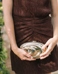 A close-up of a person wearing a brown ruched pleated dress and holding a metallic gold spiral-shaped clutch bag.