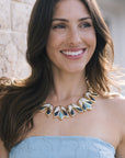 A smiling woman with long brown hair wears a chunky geometric statement necklace with gold-tone metal and light blue inlay, paired with a light blue strapless top.