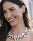 A close-up lifestyle shot of a woman wearing a geometric gold statement necklace with blue and white inlay, and matching blue stud earrings.