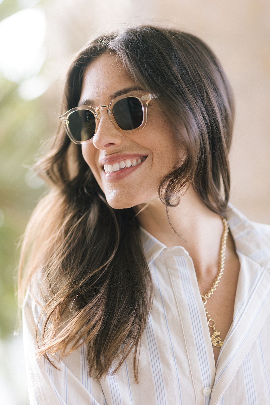 A smiling woman with long brown hair wearing round sunglasses with a clear frame and dark lenses, paired with a white striped shirt and a gold necklace.