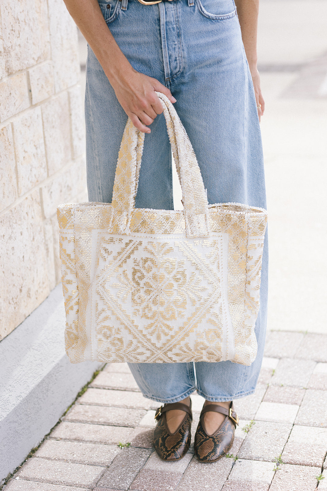 A person wearing light blue jeans holds a large cream and gold patterned tote bag with woven handles, standing on a paved sidewalk next to a light stone wall.