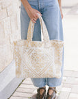 A person wearing light blue jeans holds a large cream and gold patterned tote bag with woven handles, standing on a paved sidewalk next to a light stone wall.