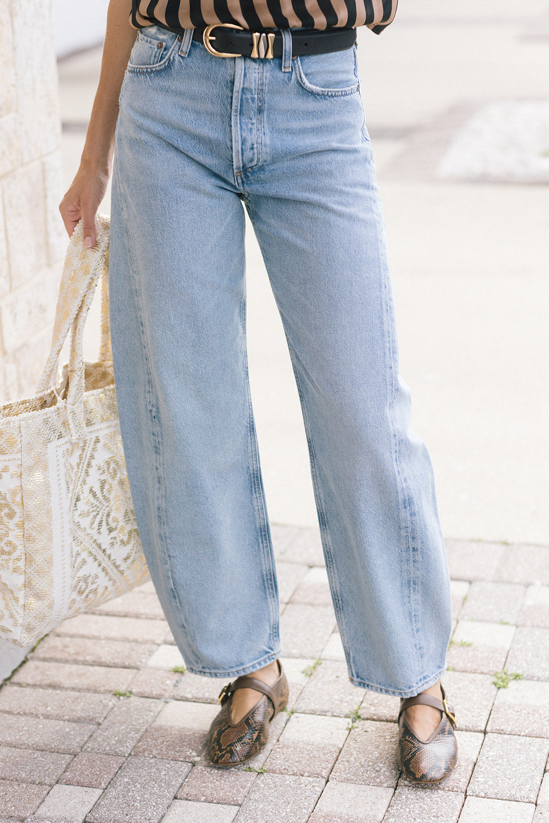 A person wearing light-wash wide-leg blue jeans with a black belt, paired with brown shoes and holding a white patterned tote bag, standing on a paved outdoor walkway.