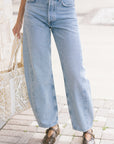 A person wearing light-wash wide-leg blue jeans with a black belt, paired with brown shoes and holding a white patterned tote bag, standing on a paved outdoor walkway.