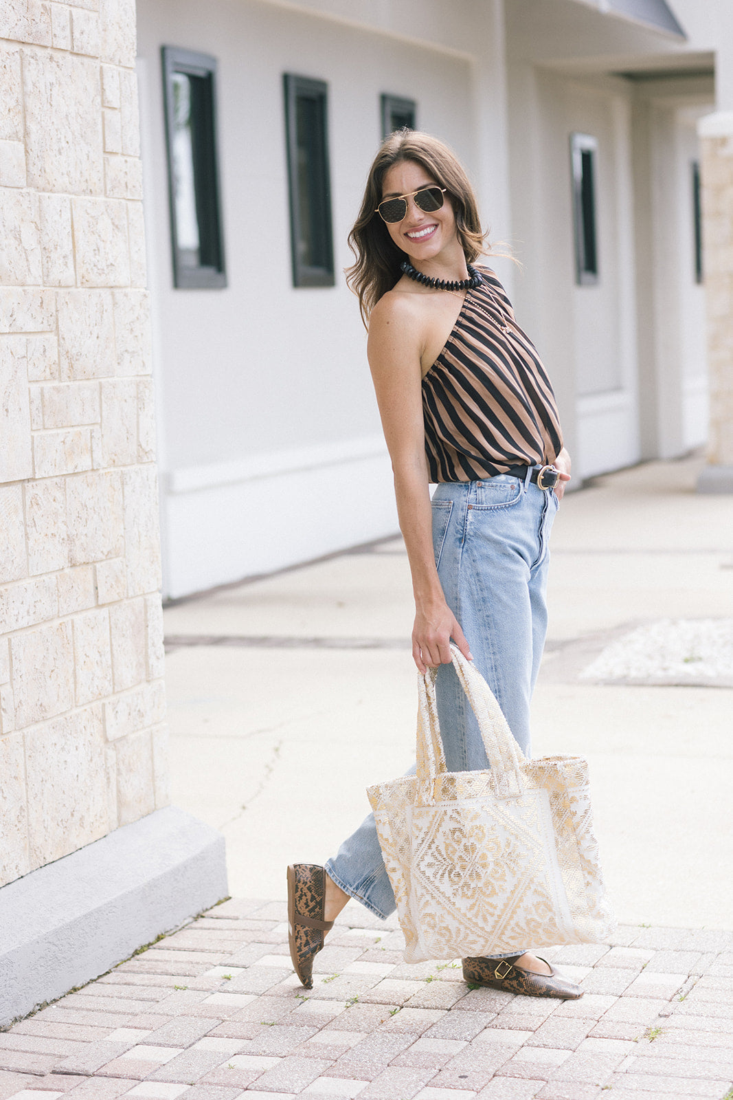A woman wearing a brown and black striped top, light blue jeans, and brown boots is walking outdoors while holding a cream and gold tote bag.