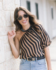 A woman wearing a black and tan striped one-shoulder blouse, light blue jeans, and sunglasses, leaning against a light beige stone wall.