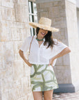 A woman wearing a white short-sleeve blouse and a green and white leaf-patterned mini skirt, accessorized with a wide-brimmed straw hat and a green beaded necklace, posing outdoors against a beige stone wall.