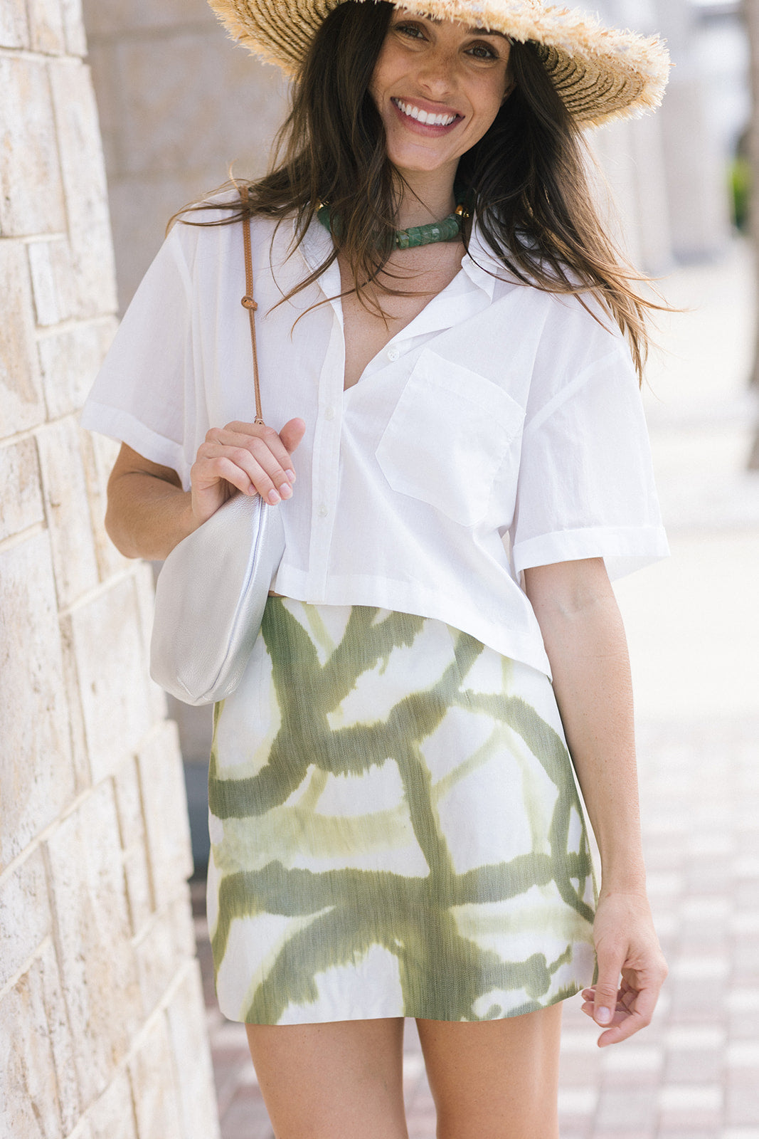 A woman wearing a white short-sleeve button-up shirt and a white mini skirt with a green print, accessorized with a straw hat, a green choker, and a white shoulder bag, standing against a light stone wall.