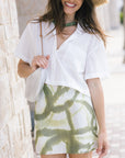 A woman wearing a white short-sleeve button-up shirt and a white mini skirt with a green print, accessorized with a straw hat, a green choker, and a white shoulder bag, standing against a light stone wall.