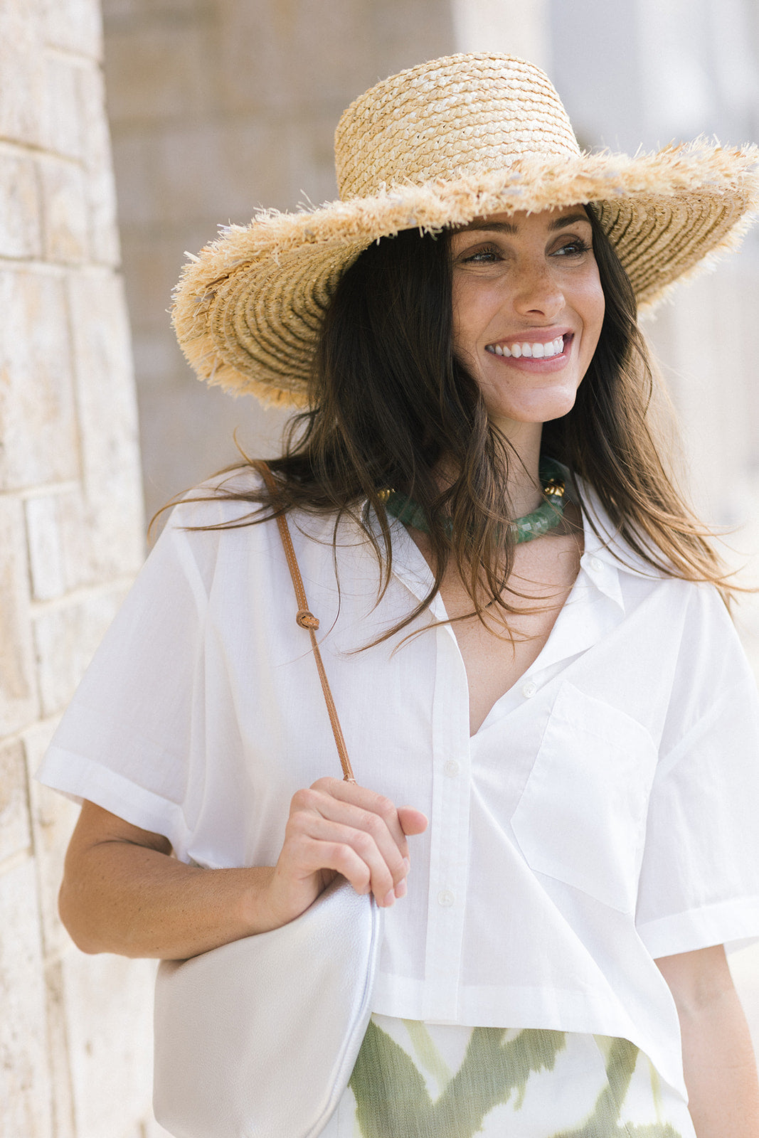 A woman wearing a wide-brimmed straw hat, a white short-sleeve button-up shirt, and a green patterned skirt, holding a white shoulder bag against a light stone wall background.