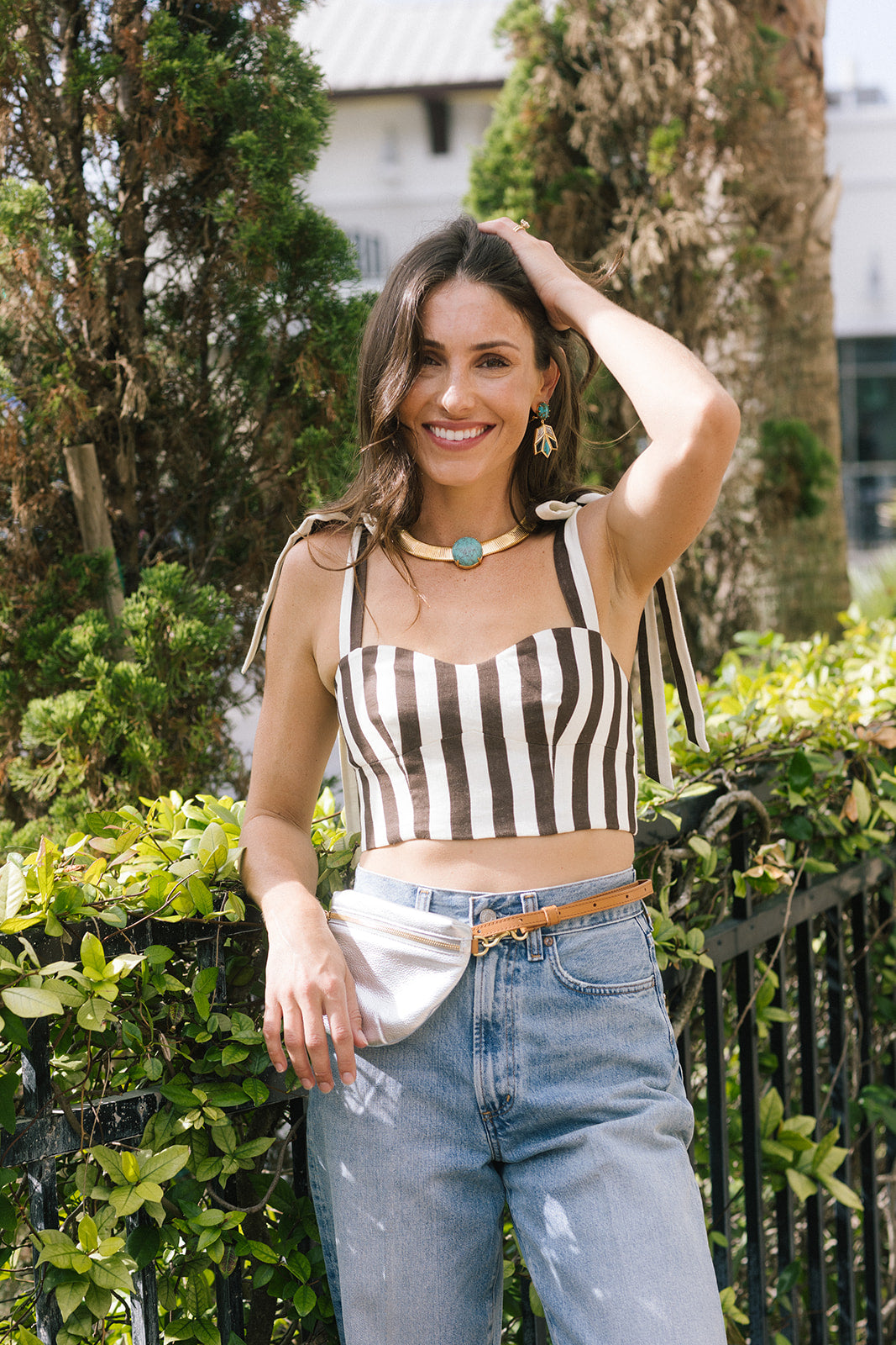 A woman posing outdoors in front of green foliage and a black metal fence, wearing a black and white vertically striped crop top with shoulder tie details, light-wash blue jeans, a silver fanny pack, and a brown belt.