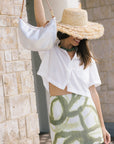 A woman wearing a white cropped button-up shirt and a white mini skirt with a green abstract pattern, styled with a wide-brimmed straw hat and a silver shoulder bag, posing against a beige stone wall.